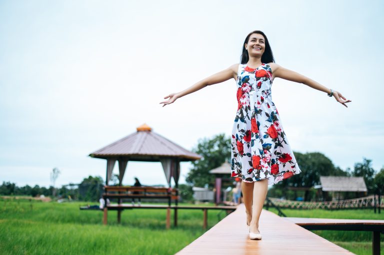 A woman in a floral dress walks barefoot on a wooden path with arms outstretched, smiling, enjoying a digital detox with a gazebo and green field in the background.