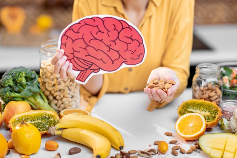 Person holding a paper brain illustration and almonds, surrounded by fruits, vegetables, and nuts on a table—highlighting how diet supports brain health and mental health.