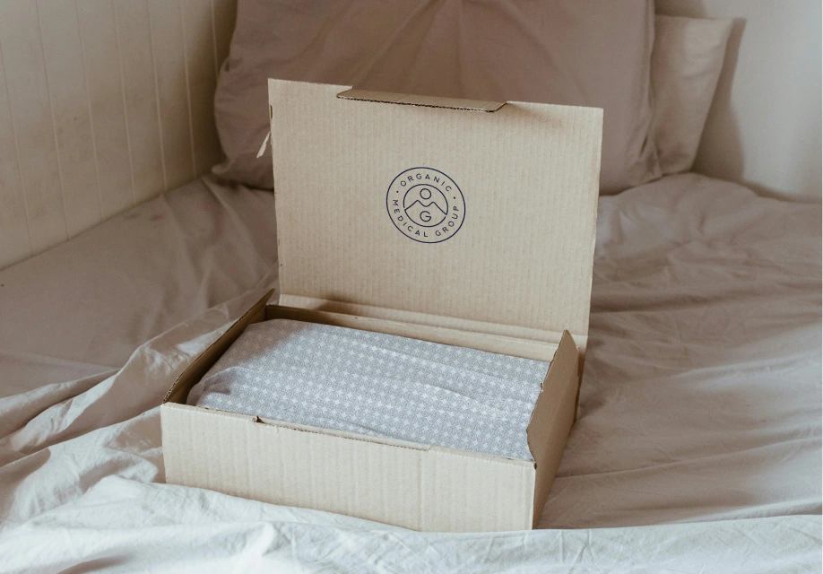 A cardboard box with a patterned fabric inside sits open on a bed with beige sheets and pillows.