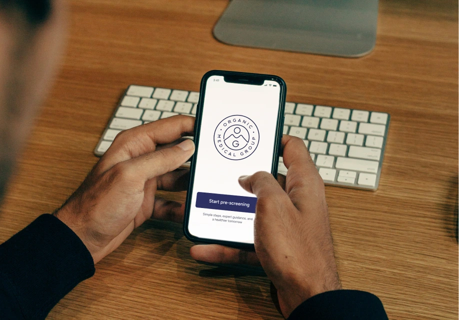 A person holds a smartphone displaying a medical group app with a “Start pre-screening” button, in front of a wireless keyboard and computer monitor on a wooden desk.