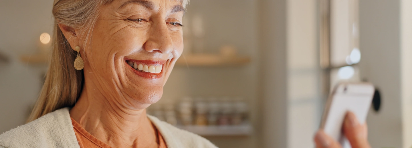 An older woman with gray hair smiles while looking at a smartphone indoors, wearing earrings and a light-colored top.