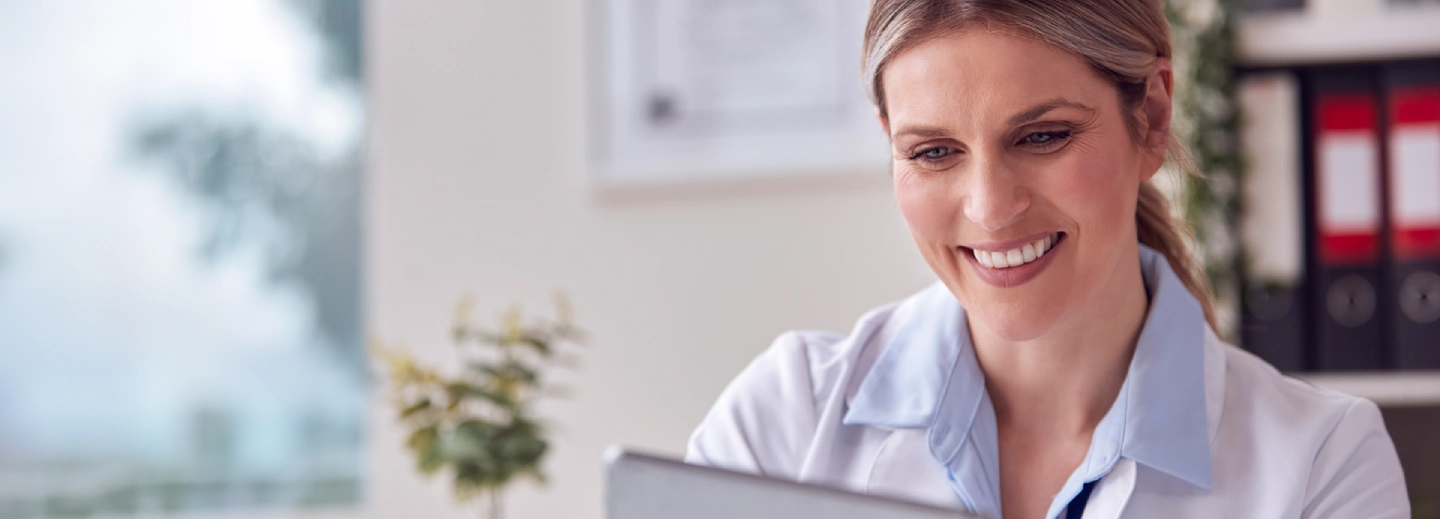A smiling woman in a white lab coat sits at a desk, looking at a laptop in a bright office setting.