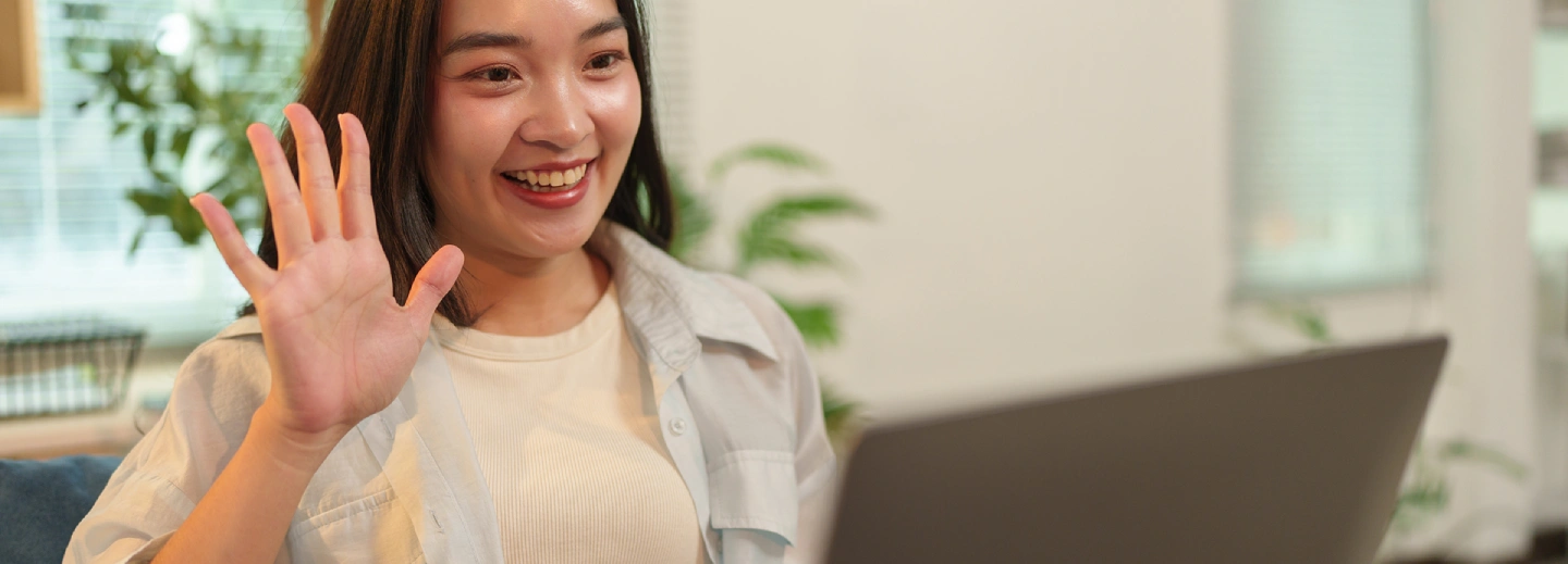 A woman sits indoors, smiling and waving at a laptop screen during a video call.