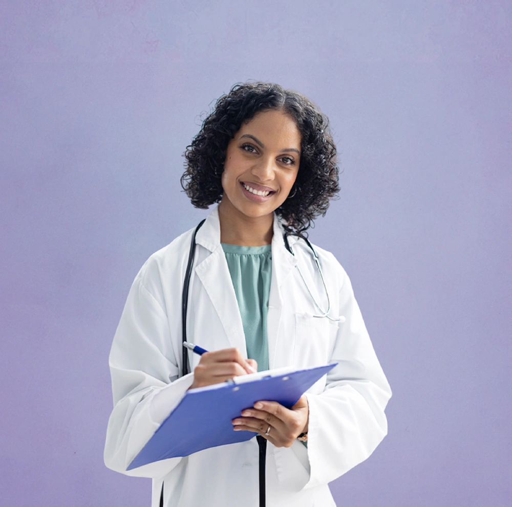 A doctor wearing a white coat and stethoscope writes on a blue clipboard while smiling against a plain purple background.