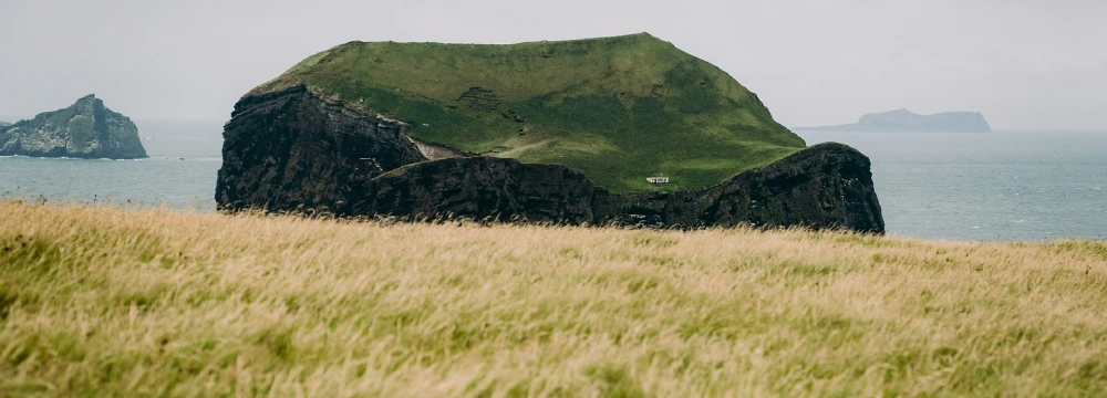 A large, grass-covered rocky island sits in the ocean, viewed from a field of tall grass with other islands visible in the distance under an overcast sky.