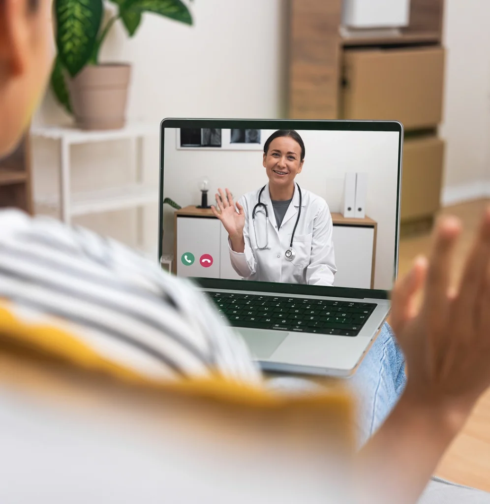 A person has a video call with a doctor, who is smiling and waving on the laptop screen.