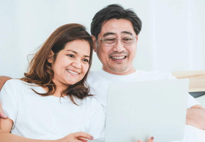 A smiling couple sits closely together on a couch, looking at a laptop screen. Both are wearing white shirts and appear relaxed.