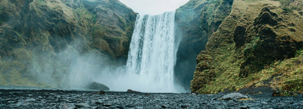 A wide waterfall cascades down a cliff between moss-covered rocks, with mist rising above a rocky ground in the foreground.